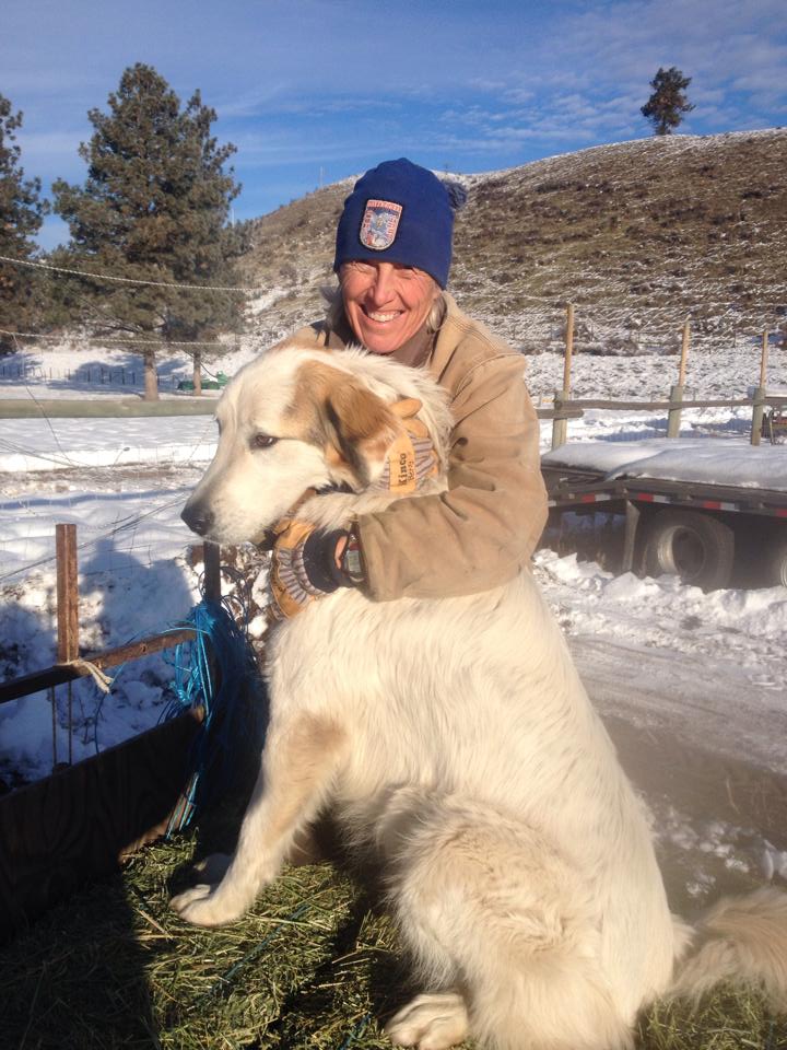 Smiling person in winter clothing hugging a large white dog outdoors in a snowy landscape, with trees and a hillside in the background.