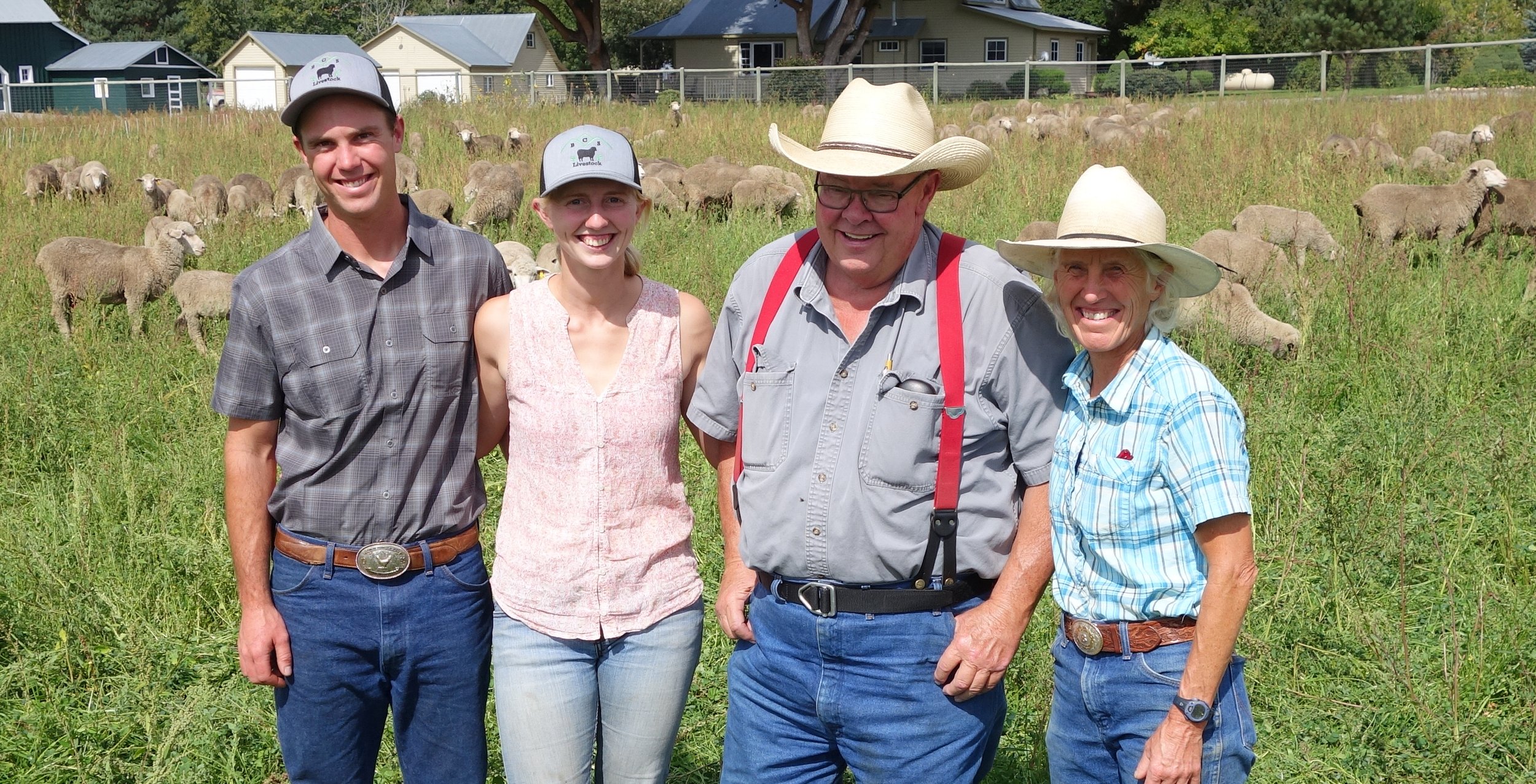 Group of four farmers posing in a lush green field with grazing sheep in the background, showcasing sustainable farming practices. The individuals are dressed in casual, farm-appropriate attire, including hats and denim, representing a family-run agricultural operation.
