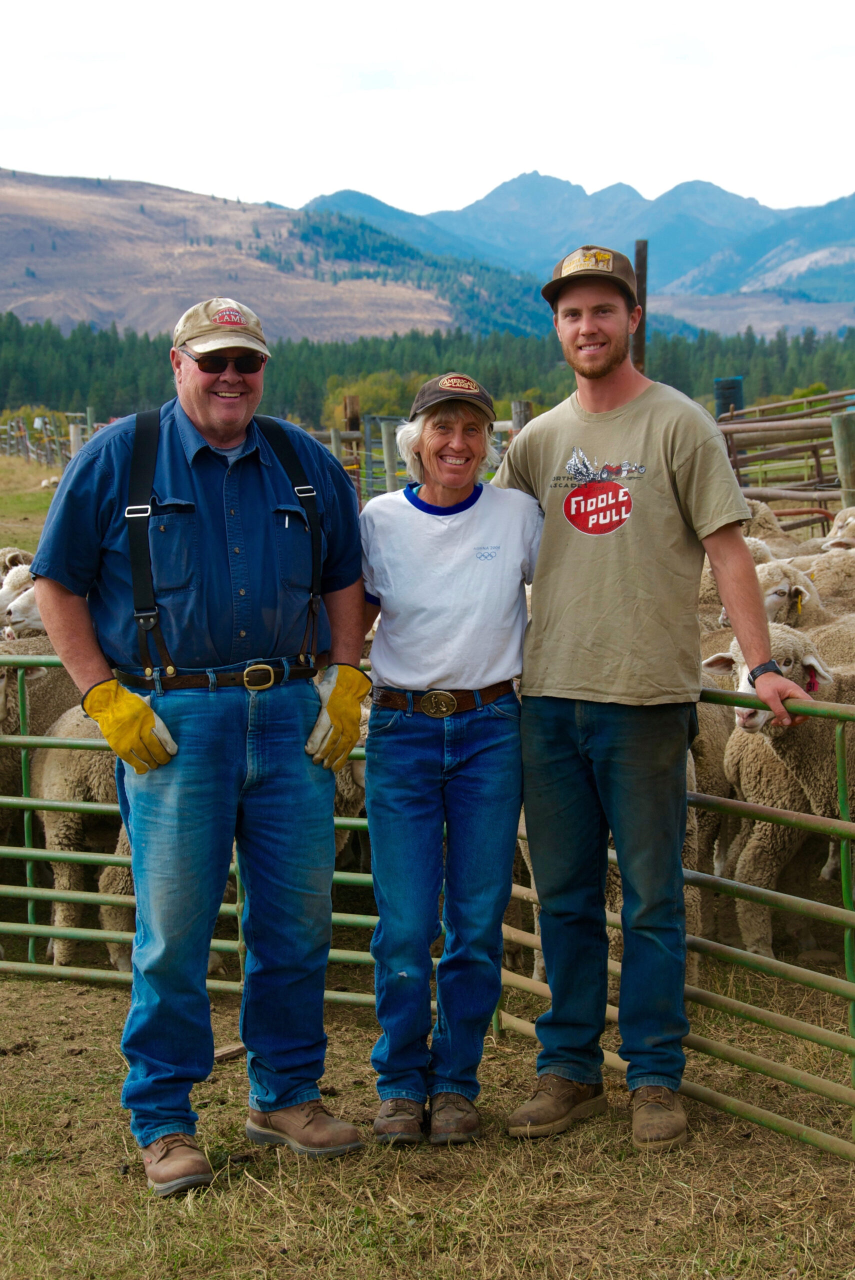 Three farmers pose together in front of sheep on a ranch, with mountains in the background. The group consists of a man wearing glasses and gloves, a woman in a white shirt, and a young man in a tan shirt. The image captures a moment of camaraderie in a rural farming setting.