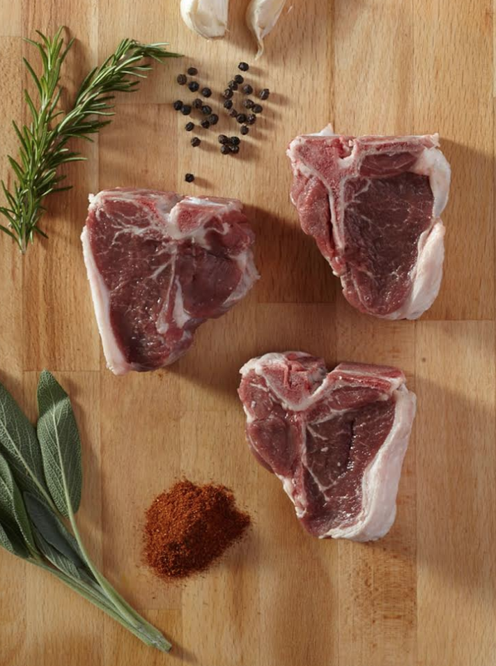 Raw lamb chops on a wooden cutting board, accompanied by fresh rosemary, sage leaves, garlic cloves, black peppercorns, and a sprinkle of red spice. Ideal for cooking enthusiasts and recipe creators.