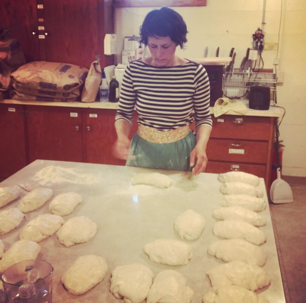 A woman in a striped shirt and apron kneads dough on a floured countertop in a bakery, surrounded by neatly shaped loaves of bread. Bags of flour are stacked in the background, highlighting a cozy baking environment.