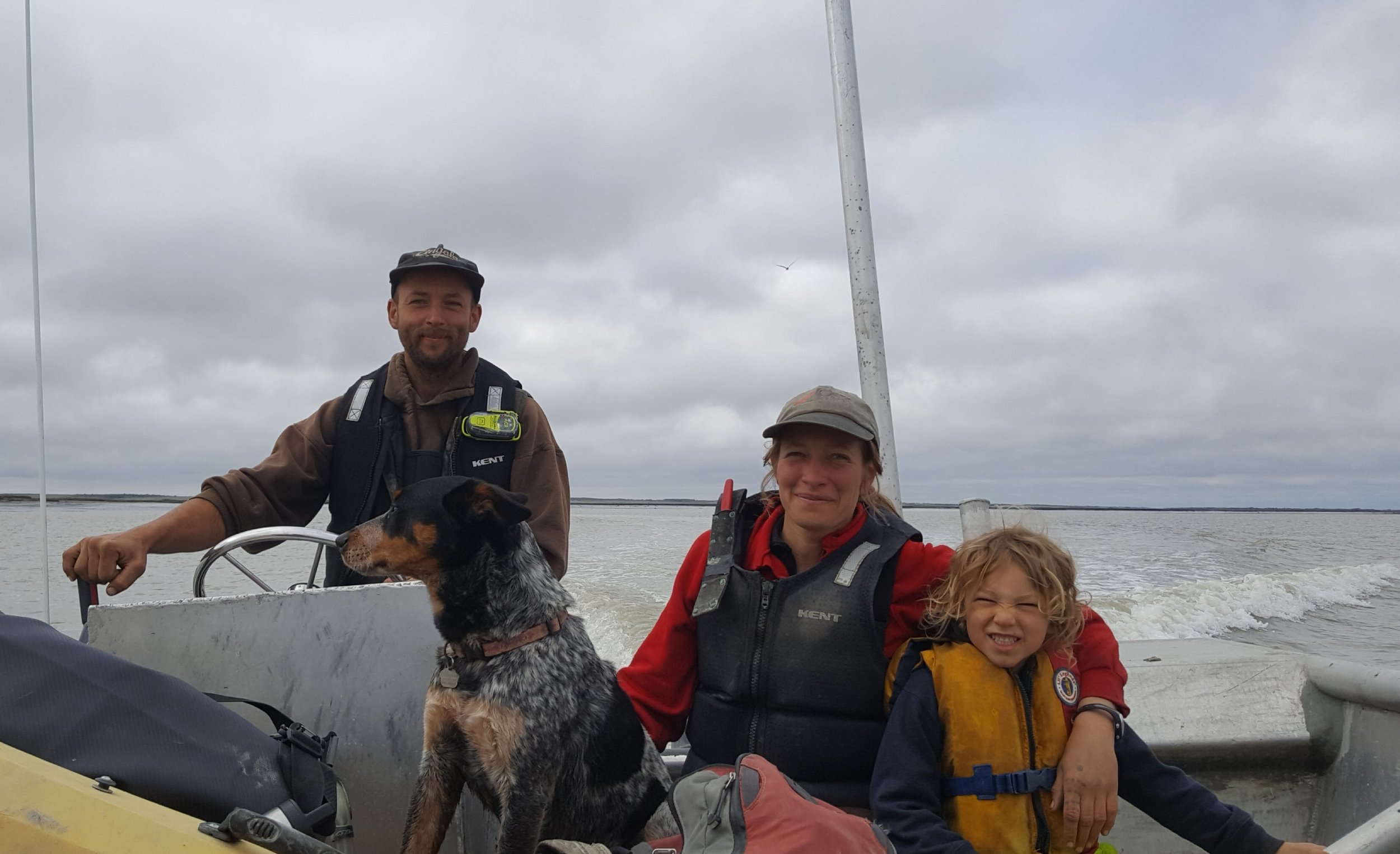 Family boating adventure with a dog, featuring a man, woman, and child wearing life jackets on a small boat in a cloudy outdoor setting.