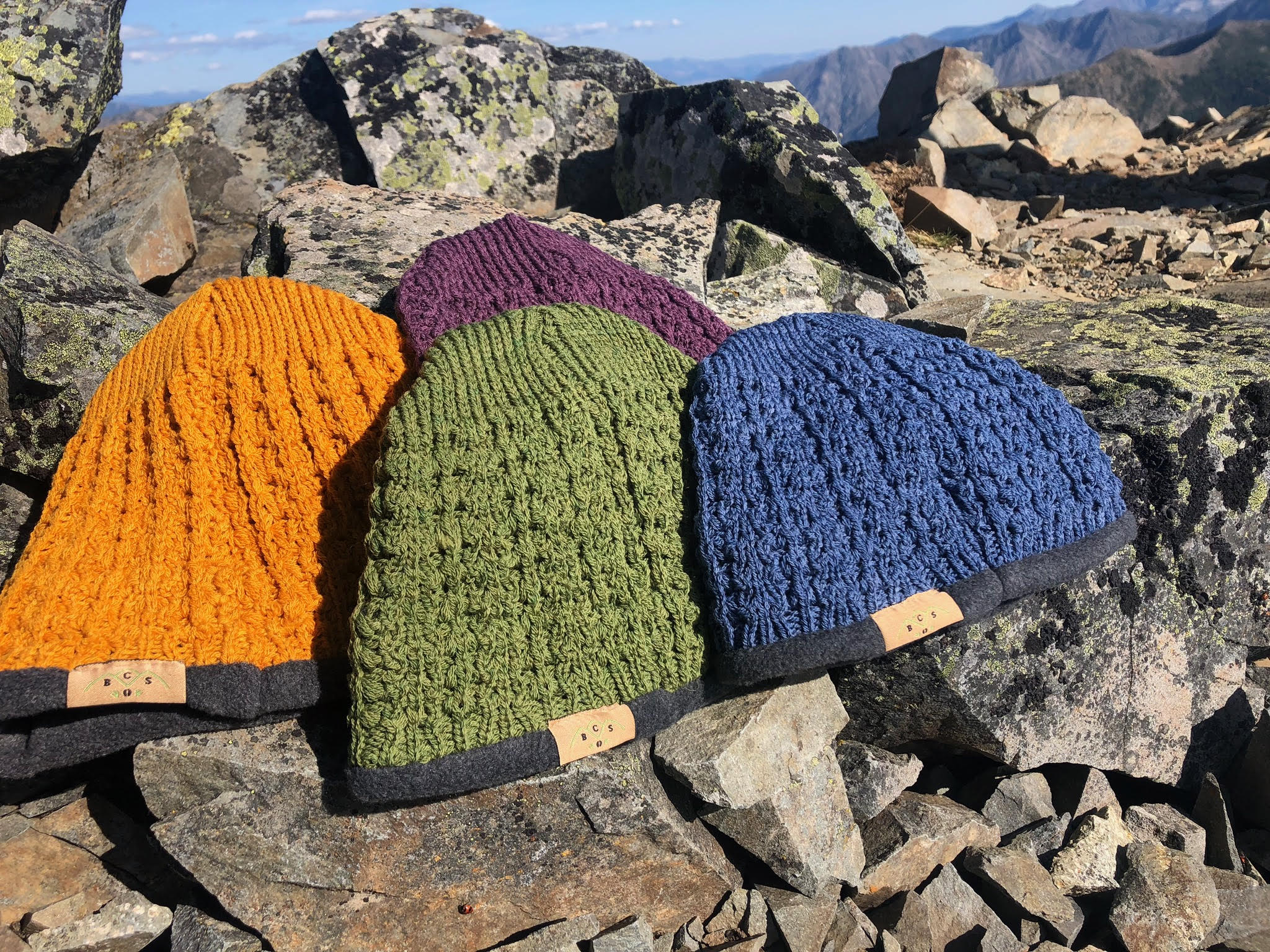 Colorful knitted beanies in orange, green, purple, and blue displayed on rocky terrain, showcasing their textured patterns and stylish design, ideal for outdoor winter activities.